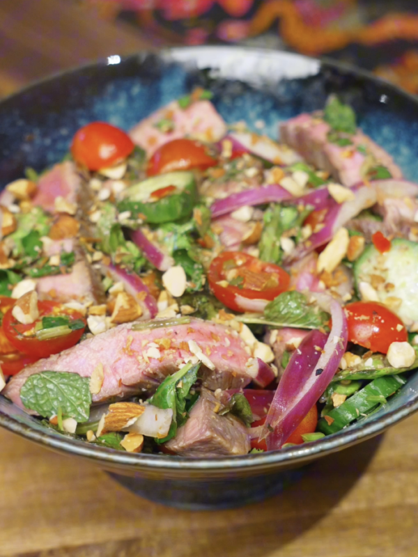 Finished Thai beef salad served in a bowl with sliced steak, red onion, cucumber, cherry tomatoes, fresh herbs, and chopped nuts tossed in a lime-forward dressing.