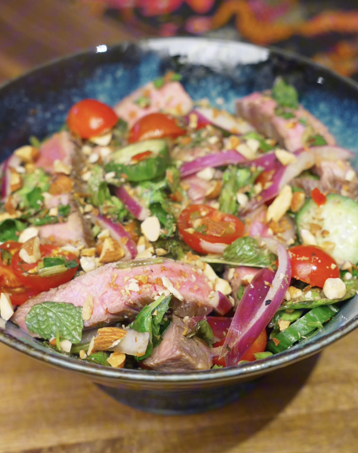 Finished Thai beef salad served in a bowl with sliced steak, red onion, cucumber, cherry tomatoes, fresh herbs, and chopped nuts tossed in a lime-forward dressing.