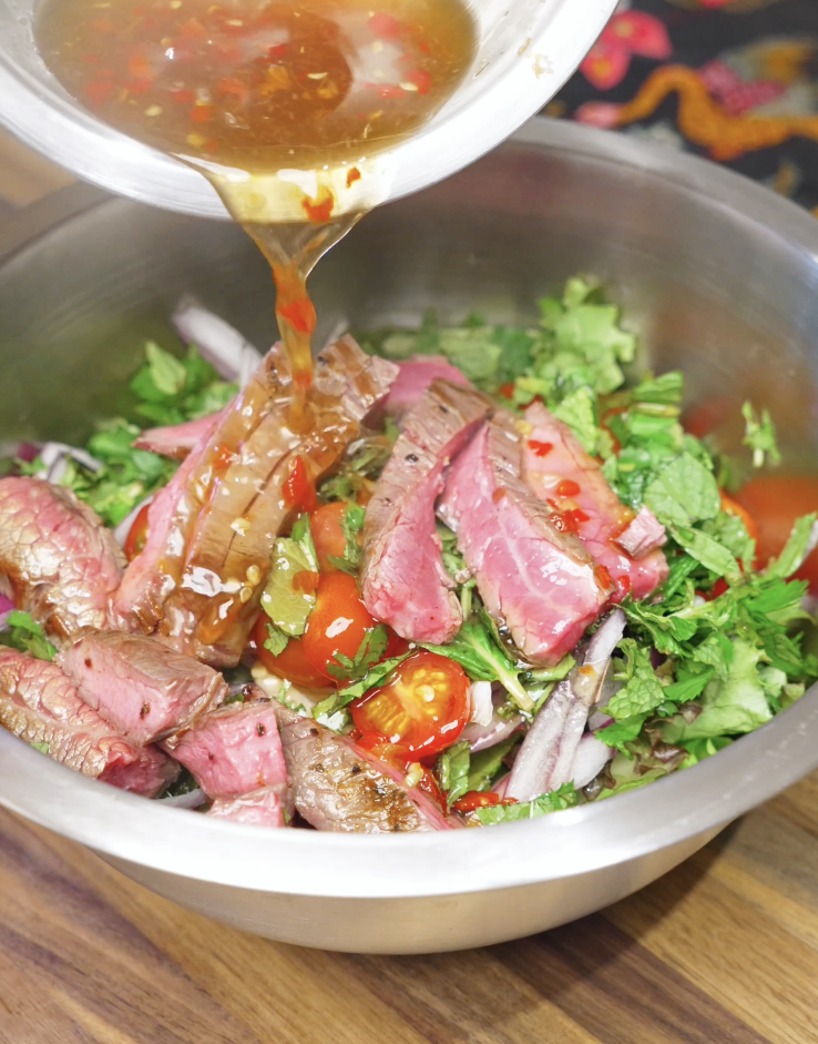 Pouring tangy Thai beef salad dressing over sliced steak, herbs, and vegetables in a mixing bowl, evenly coating everything with lime and fish sauce flavor.