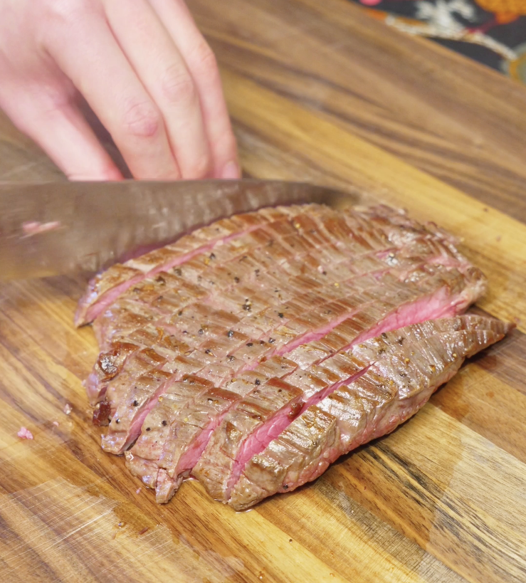 Slicing seared flank steak thinly against the grain on a wooden cutting board to ensure tender slices for Thai beef salad.
