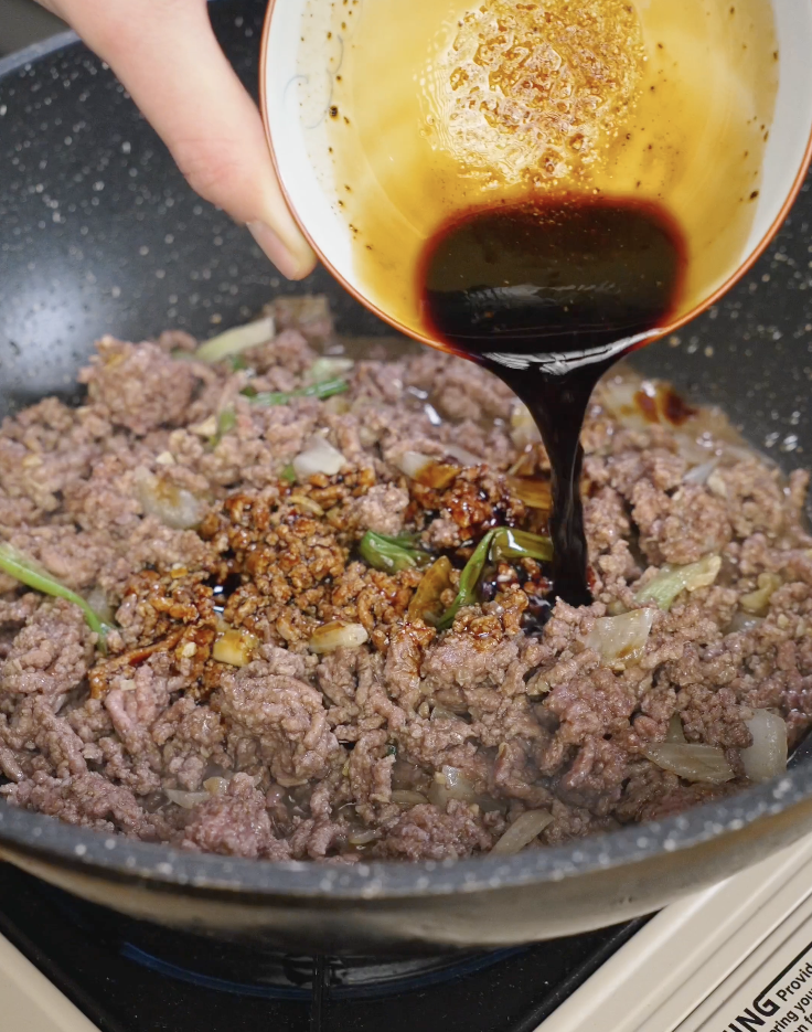 Dark soy-based black pepper sauce being poured over browned ground beef in a skillet, coating the meat with a glossy, peppery glaze.