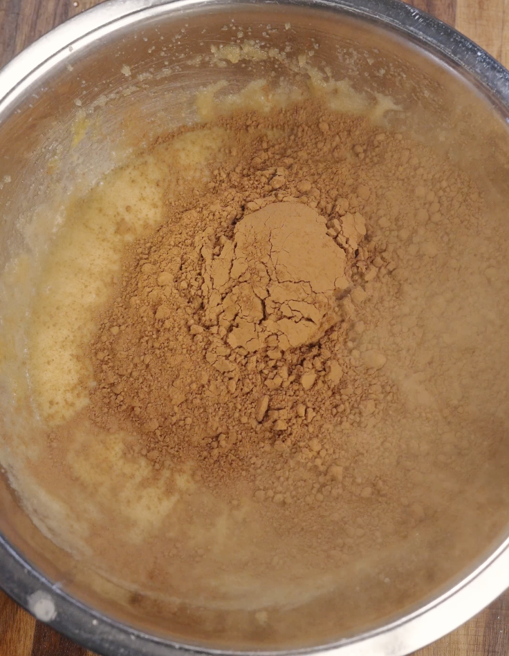 Unsweetened cocoa powder added to the brownie batter in a mixing bowl, deepening the chocolate flavor for fudgy Oreo brownies.