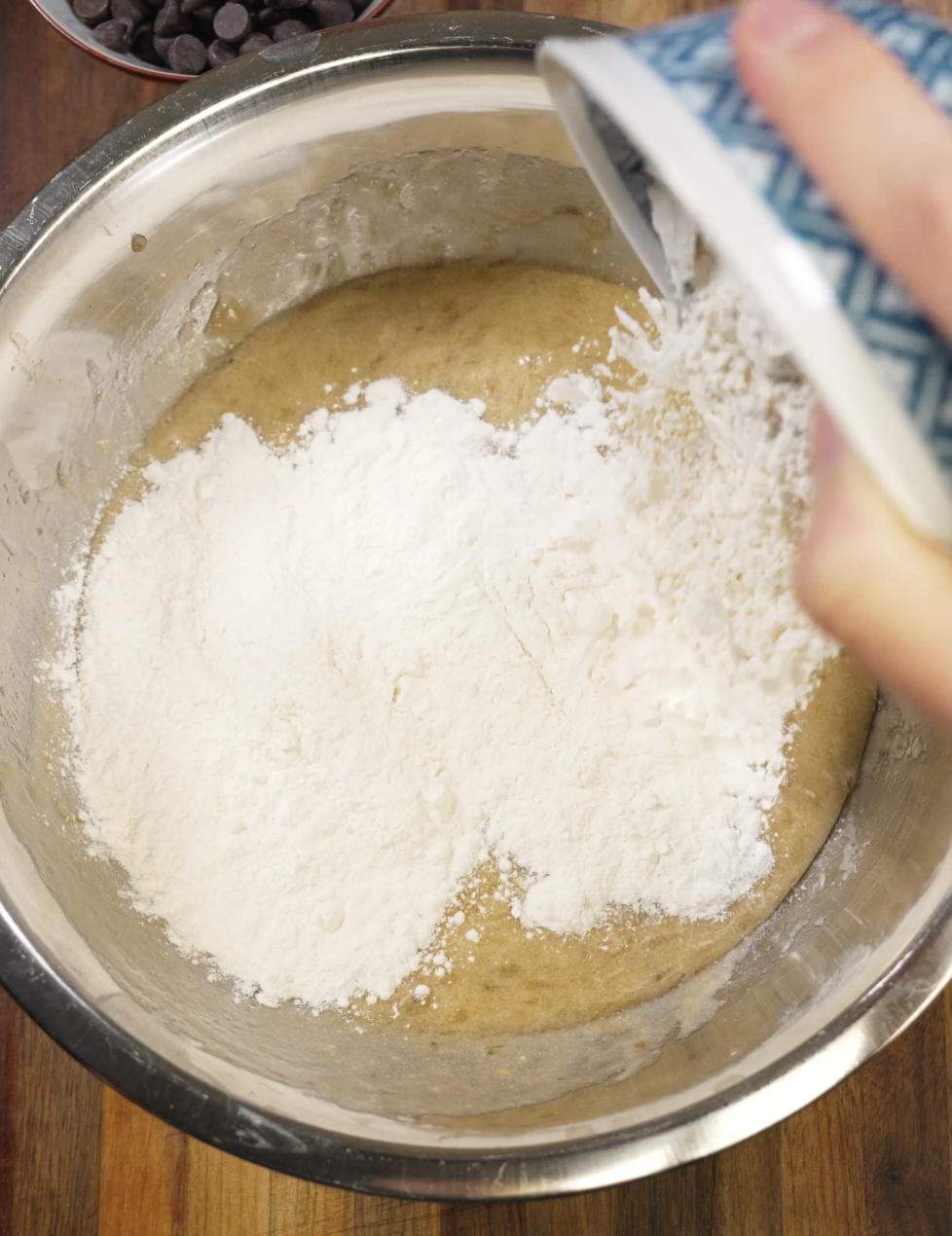 All-purpose flour being added to a bowl of banana bread batter made with mashed ripe bananas, melted butter, brown sugar, and egg before gently folding to create a moist chocolate chip banana bread.