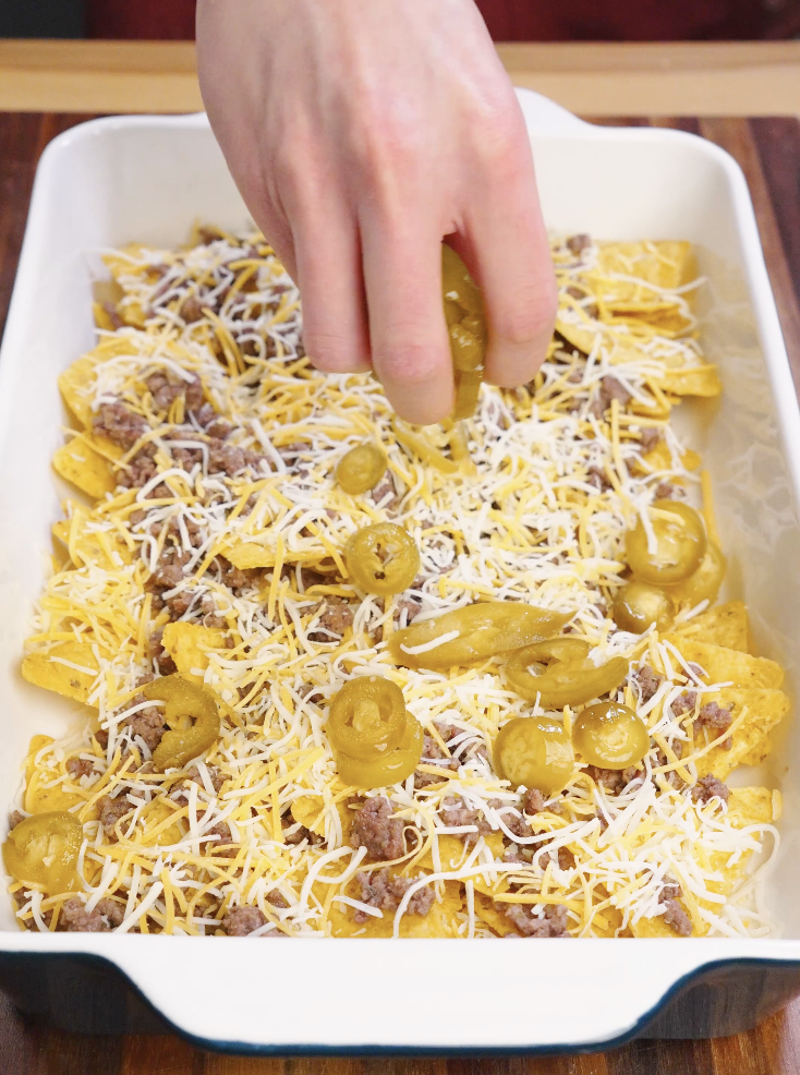 Pickled jalape&ntilde;o slices being placed over tortilla chips, seasoned ground beef, and shredded cheese while assembling sheet pan beef nachos.