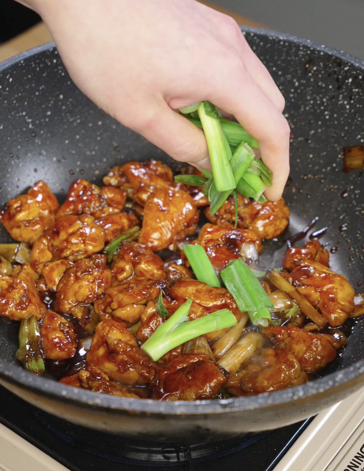 Fresh scallion greens being added to glossy Mongolian chicken in a skillet just before serving to brighten the sweet savory sauce.