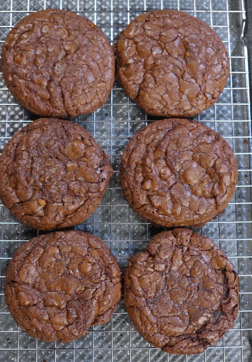 Freshly baked brownie cookies cooling on a wire rack with glossy crackled tops and set edges, showing proper sugar dissolution and aeration.