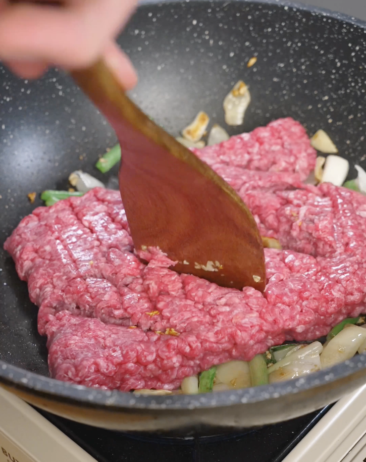 Ground beef being broken apart with a wooden spatula in a skillet with saut&eacute;ed garlic and onions while cooking for black pepper ground beef.