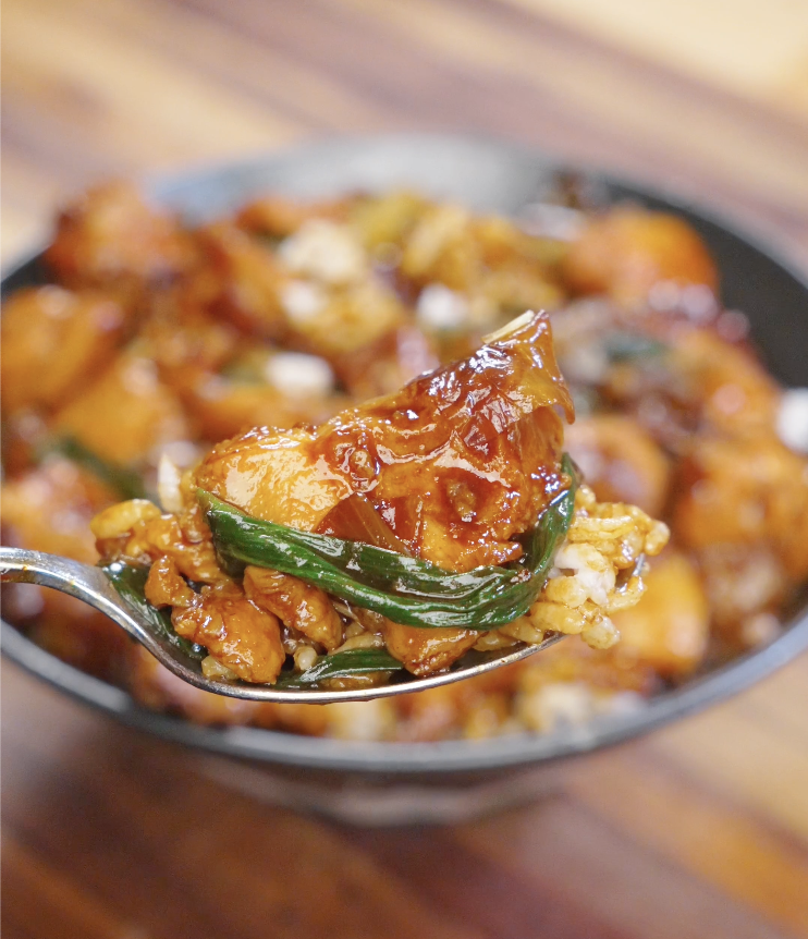 Spoonful of tender chicken, onion, and scallions coated in savory stir fry sauce lifted over a bowl of steamed rice.