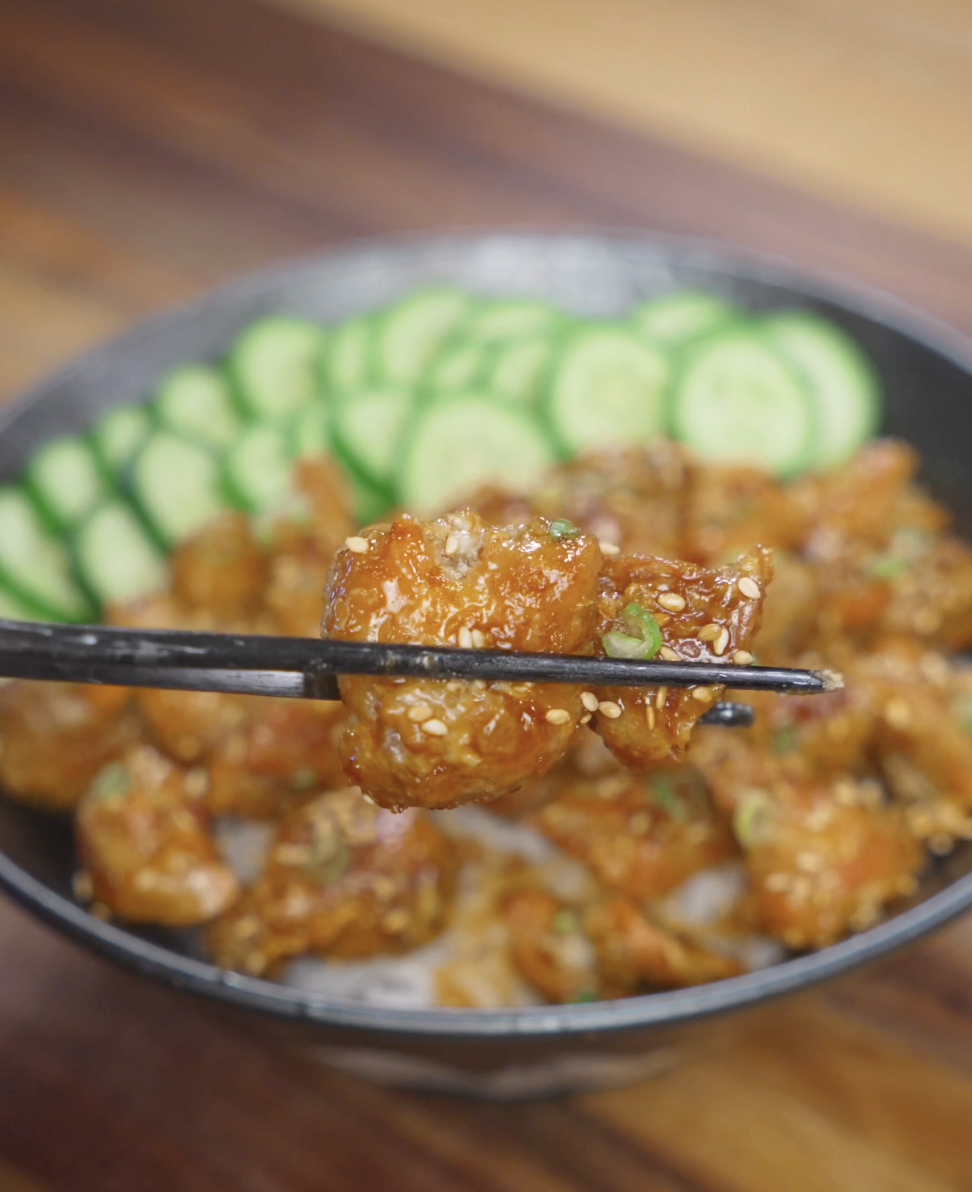 Close-up of a single piece of crispy Korean chicken held with chopsticks, showing the golden, crunchy exterior coated in sticky soy garlic sauce over a rice bowl.
