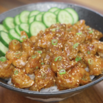 Finished bowl of crispy Korean chicken made with air-fried cornstarch-coated chicken thighs tossed in a glossy soy garlic sauce, served over steamed rice with sliced cucumbers, sesame seeds, and scallions.