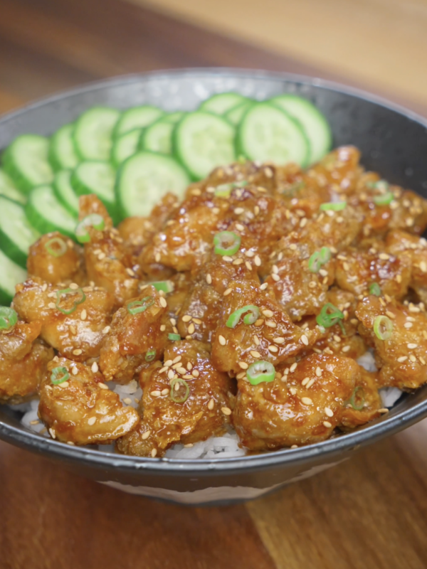 Finished bowl of crispy Korean chicken made with air-fried cornstarch-coated chicken thighs tossed in a glossy soy garlic sauce, served over steamed rice with sliced cucumbers, sesame seeds, and scallions.