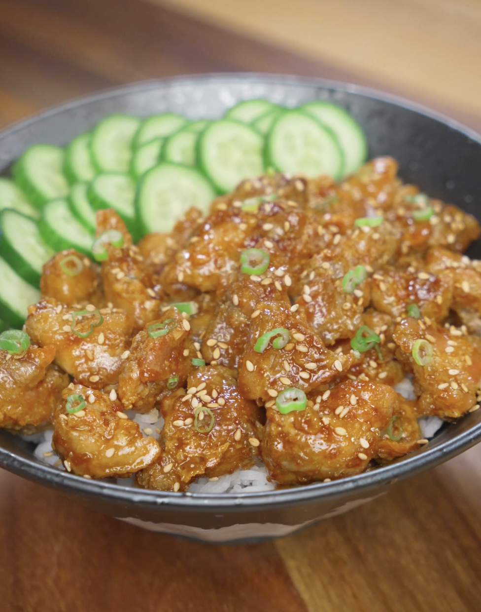 Finished bowl of crispy Korean chicken made with air-fried cornstarch-coated chicken thighs tossed in a glossy soy garlic sauce, served over steamed rice with sliced cucumbers, sesame seeds, and scallions.