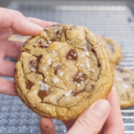 Single brown butter chocolate chip cookie held in hand, showing crisp edges, a soft center, melted chocolate chunks, and flaky salt on top.
