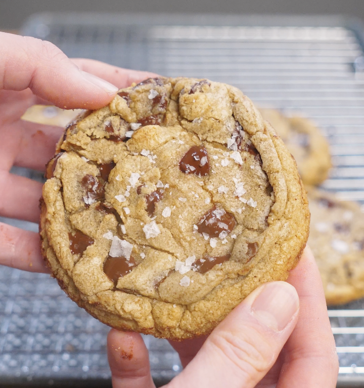 Single brown butter chocolate chip cookie held in hand, showing crisp edges, a soft center, melted chocolate chunks, and flaky salt on top.