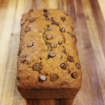 Freshly baked chocolate chip banana bread loaf with a golden brown top and melted chocolate chips, cooled on a wooden cutting board and ready to slice.