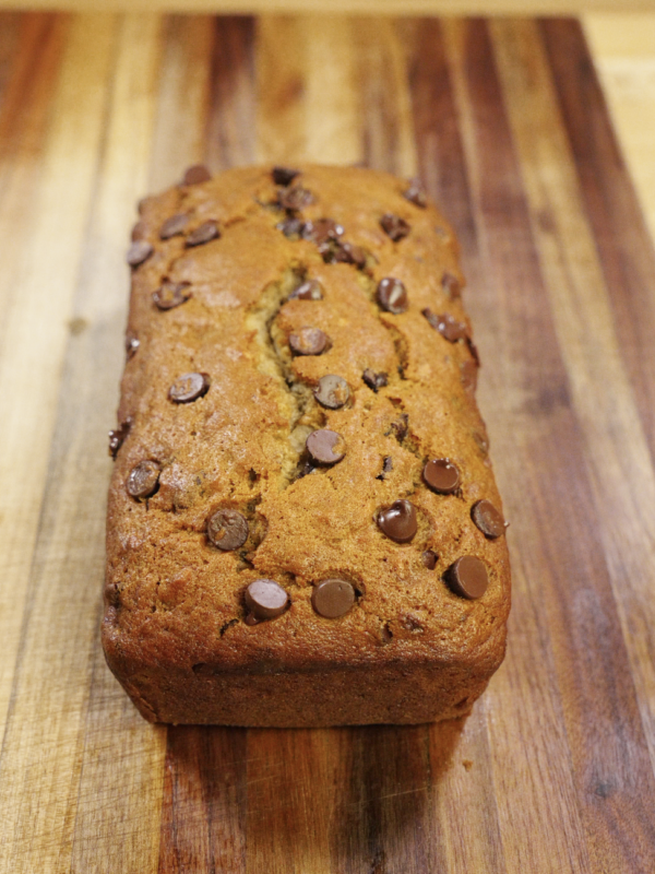 Freshly baked chocolate chip banana bread loaf with a golden brown top and melted chocolate chips, cooled on a wooden cutting board and ready to slice.
