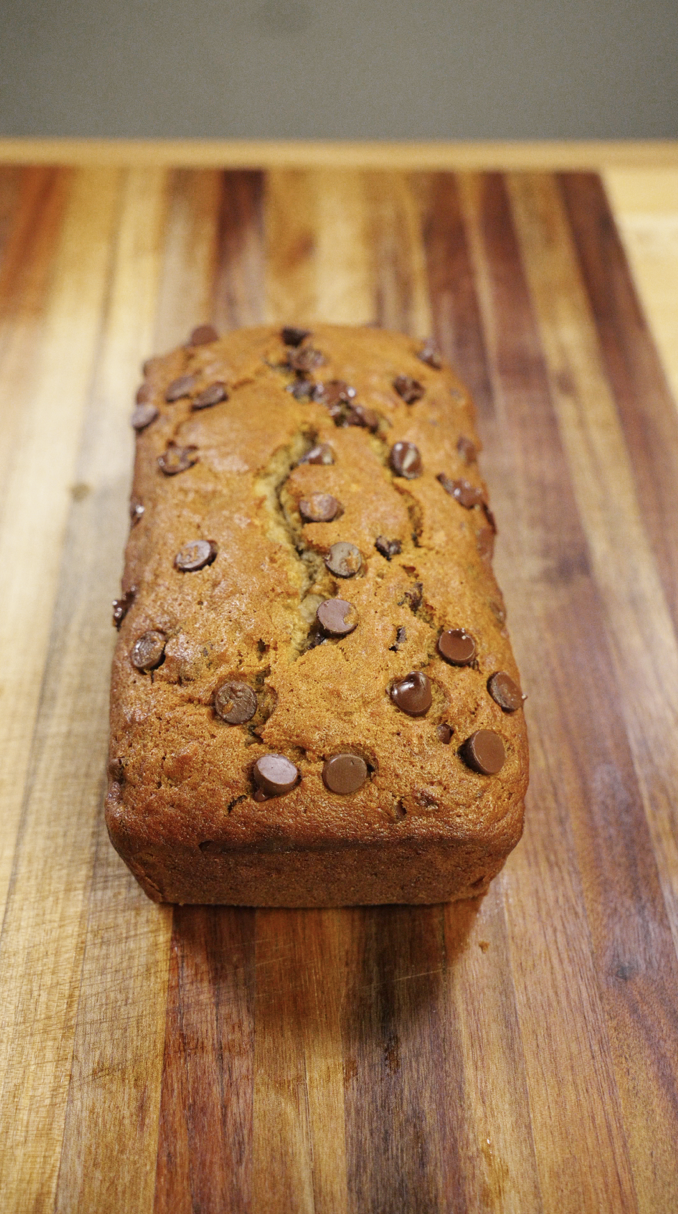 Freshly baked chocolate chip banana bread loaf with a golden brown top and melted chocolate chips, cooled on a wooden cutting board and ready to slice.