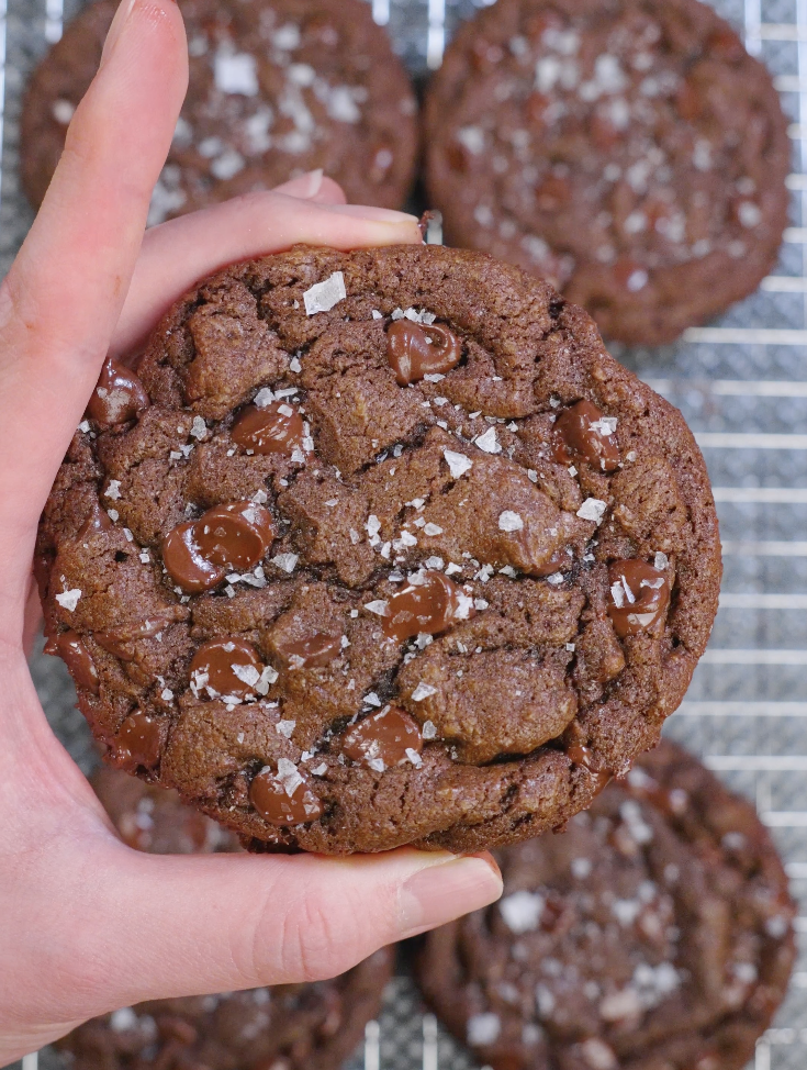 Thick double chocolate brown butter cookies baked until set with cracked tops, melted chocolate puddles, and flaky sea salt on a cooling rack.