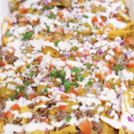 Fully assembled sheet pan beef nachos topped with melted cheese, seasoned ground beef, pickled jalapeños, chopped tomatoes, red onion, fresh cilantro, and drizzled garlic sour cream.