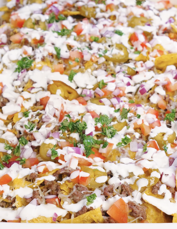 Fully assembled sheet pan beef nachos topped with melted cheese, seasoned ground beef, pickled jalapeños, chopped tomatoes, red onion, fresh cilantro, and drizzled garlic sour cream.