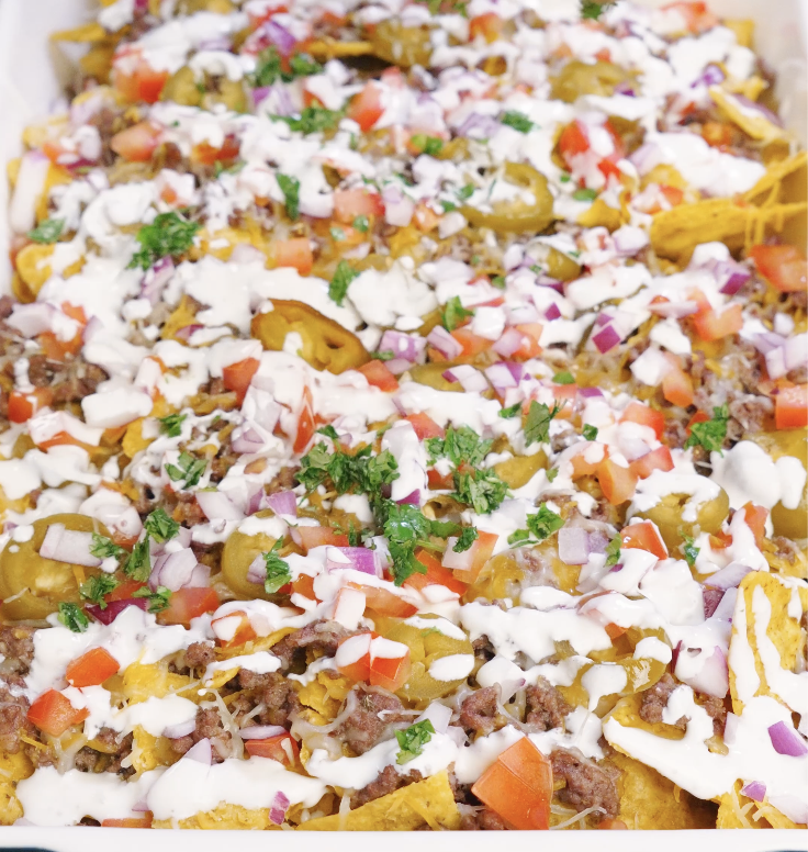 Fully assembled sheet pan beef nachos topped with melted cheese, seasoned ground beef, pickled jalape&ntilde;os, chopped tomatoes, red onion, fresh cilantro, and drizzled garlic sour cream.
