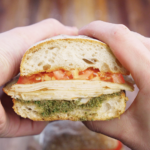 Turkey pesto sandwich cut in half and held up to show layers of seared deli turkey, melted provolone cheese, fresh tomato slices, and walnut basil pesto on toasted ciabatta bread.