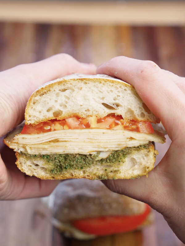 Turkey pesto sandwich cut in half and held up to show layers of seared deli turkey, melted provolone cheese, fresh tomato slices, and walnut basil pesto on toasted ciabatta bread.