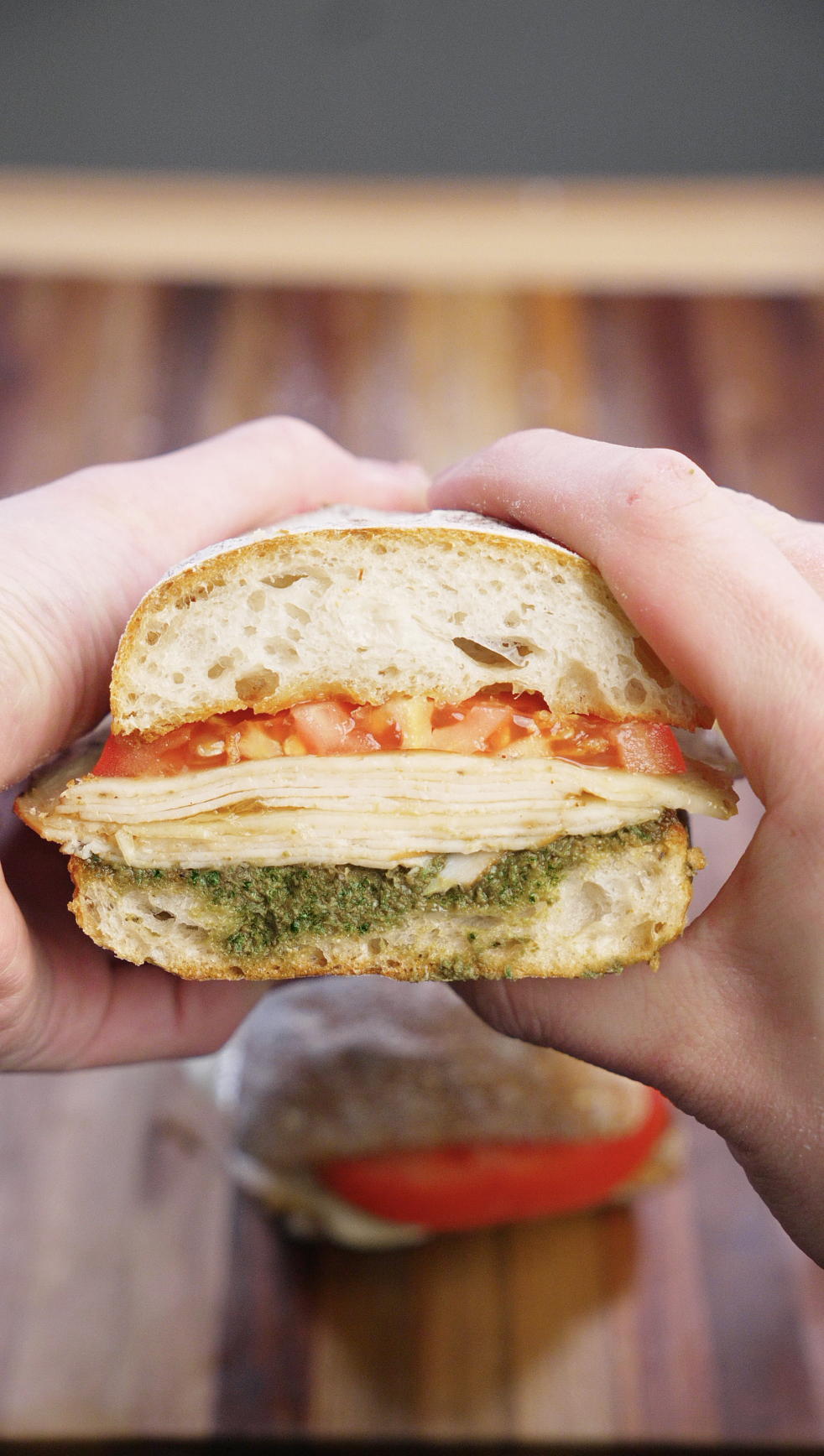 Turkey pesto sandwich cut in half and held up to show layers of seared deli turkey, melted provolone cheese, fresh tomato slices, and walnut basil pesto on toasted ciabatta bread.