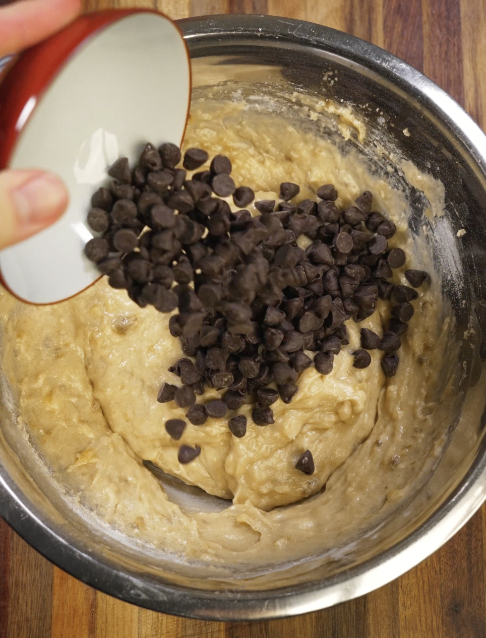 Semi-sweet chocolate chips being folded into thick banana bread batter, evenly distributing chocolate throughout the batter before baking.