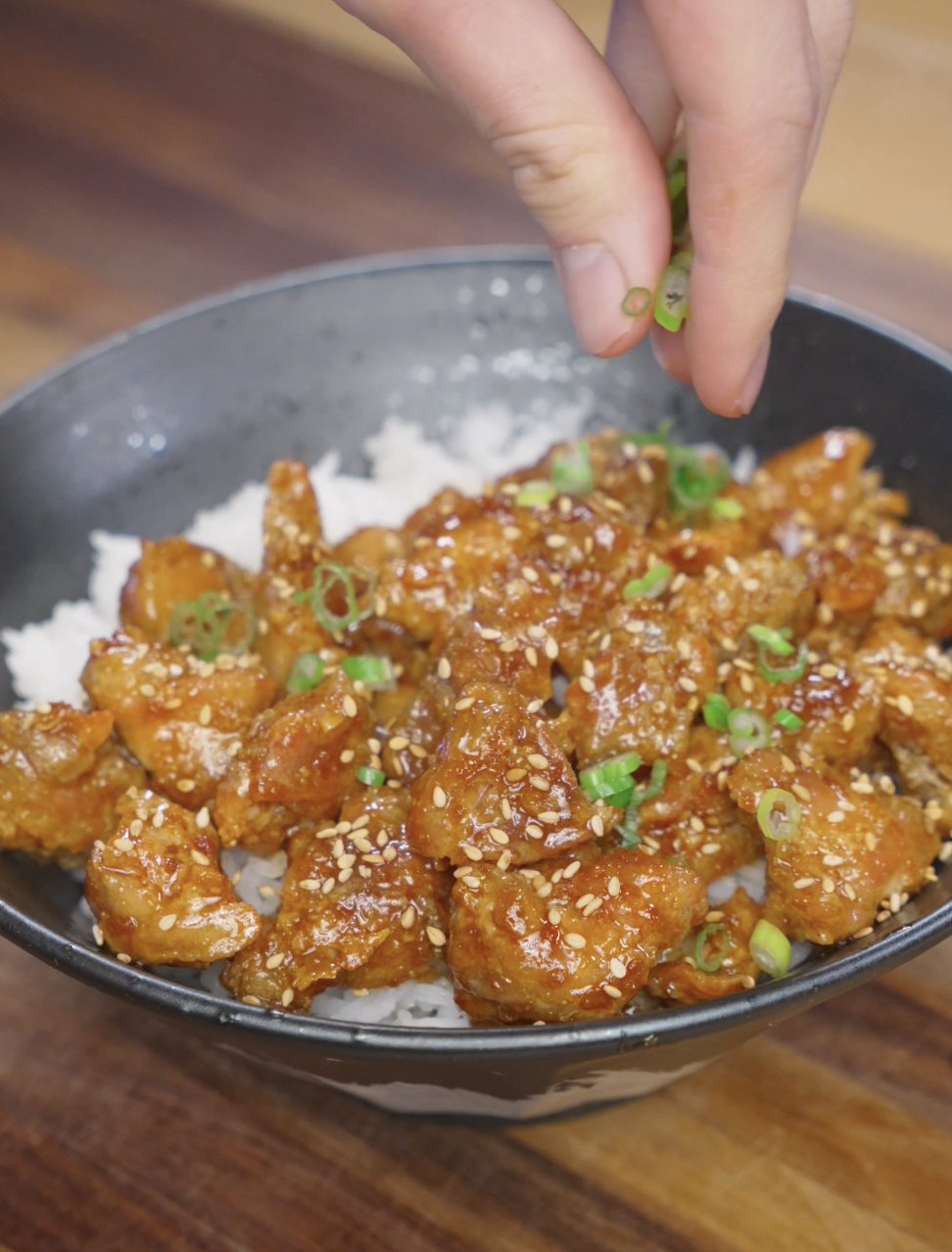 Finished crispy Korean chicken bowl being garnished with sliced scallions and sesame seeds, served over rice with cucumbers for a balanced weeknight dinner.