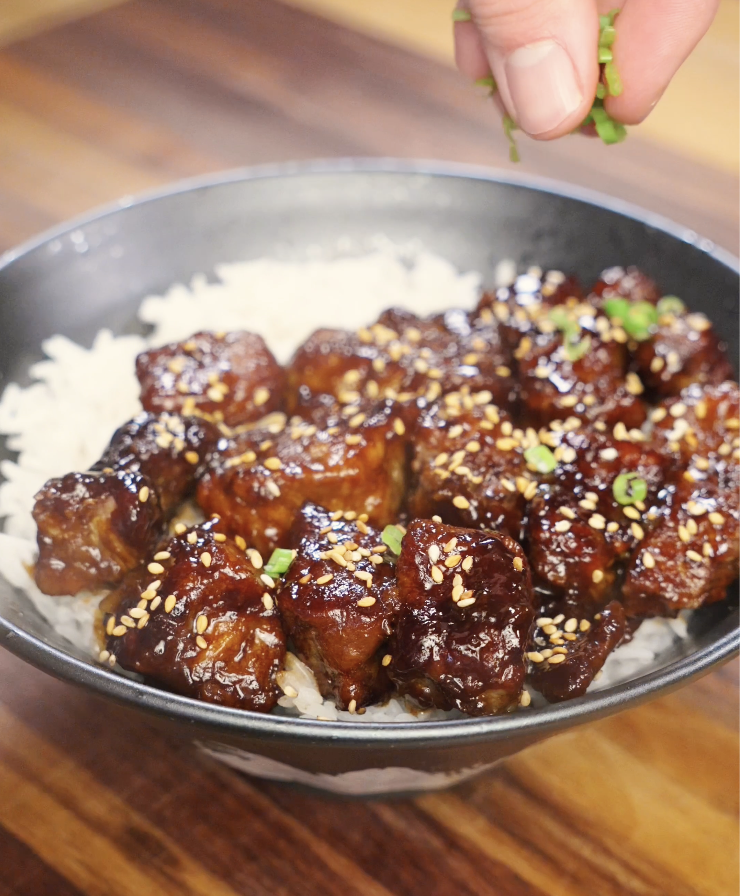 Hand sprinkling sliced scallions over soy garlic beef served on rice, adding freshness and color to the finished healthy Asian beef bowl.