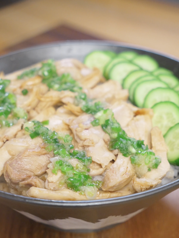 Finished bowl of ginger scallion chicken made with juicy poached chicken thighs shredded and tossed with soy sauce, topped with fragrant ginger scallion oil and served over white rice with fresh cucumber slices.