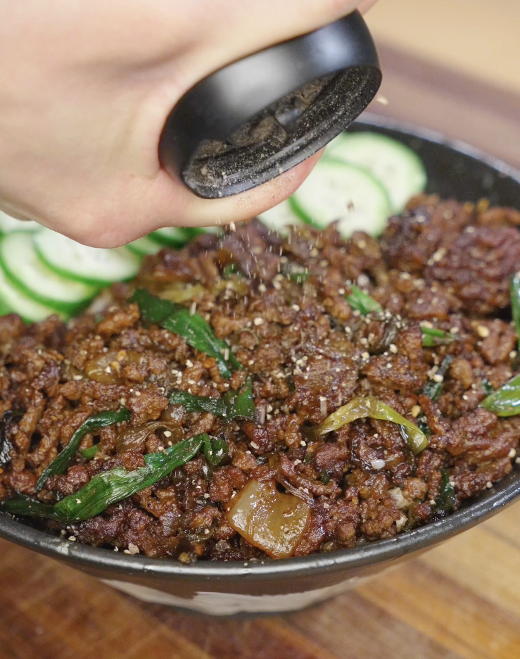 Freshly cracked black pepper being ground over a bowl of black pepper ground beef and rice to enhance aroma and spice before serving.