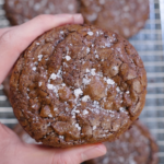 Close-up of a thick brownie cookie held in hand, showing a shiny crackled top finished with flaky sea salt and a dense, fudgy chocolate texture.