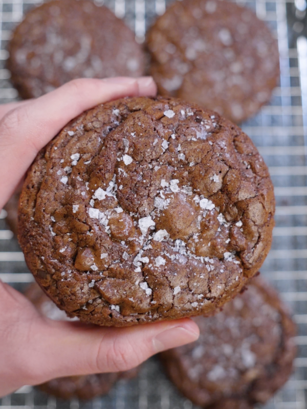 Close-up of a thick brownie cookie held in hand, showing a shiny crackled top finished with flaky sea salt and a dense, fudgy chocolate texture.