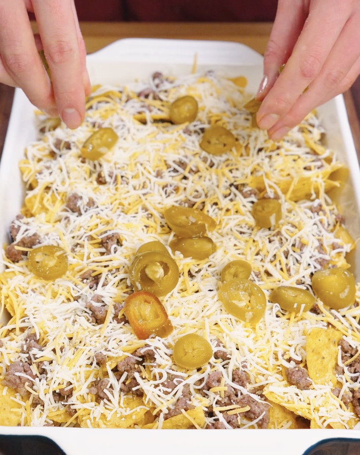 Hands sprinkling pickled jalape&ntilde;os evenly over layered tortilla chips, ground beef, and shredded cheese on a sheet pan before baking.