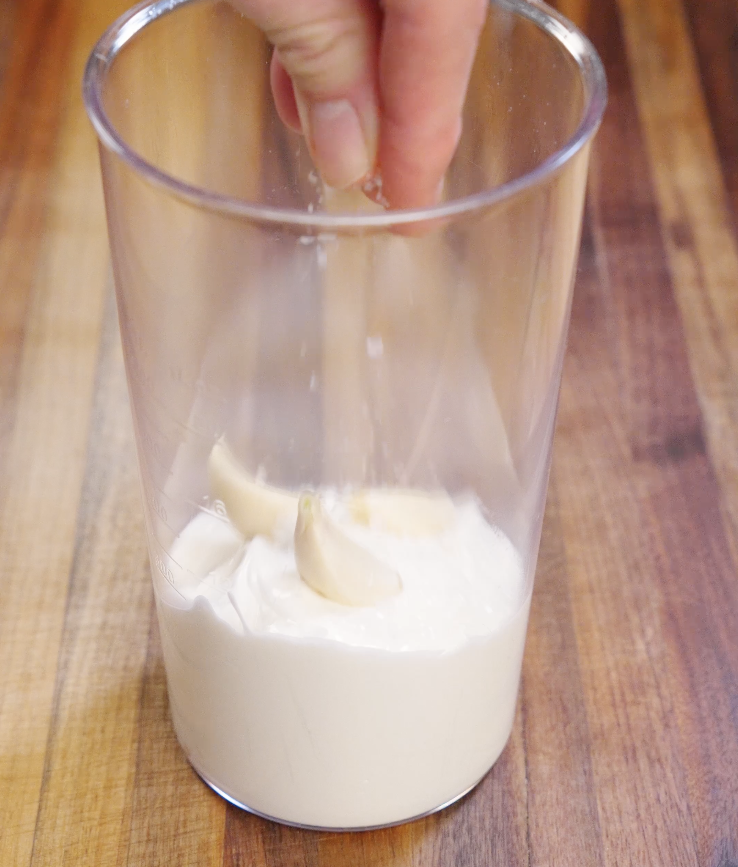 Fresh garlic being added to sour cream in a blending cup to make a smooth garlic sour cream sauce for sheet pan beef nachos.