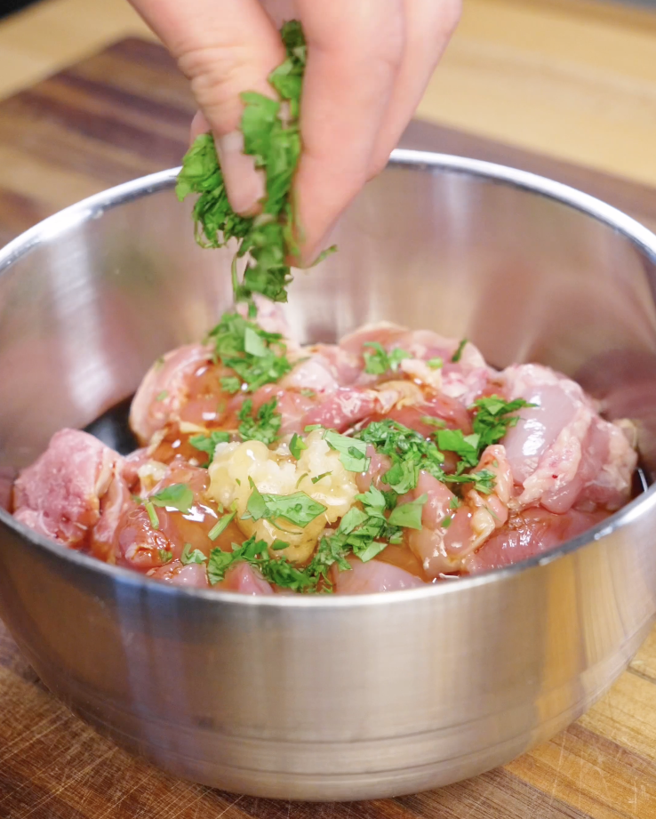 Bite sized chicken thighs being marinated in dark soy sauce, light soy sauce, vinegar, honey, garlic, and freshly chopped cilantro in a stainless steel bowl for Sticky Cilantro Chicken.