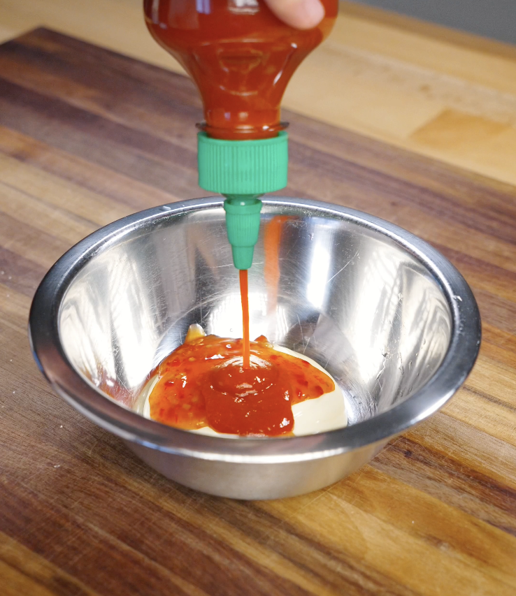 Thai sweet chili sauce and sriracha being squeezed into a bowl of mayonnaise to make homemade bang bang sauce for crispy chicken.