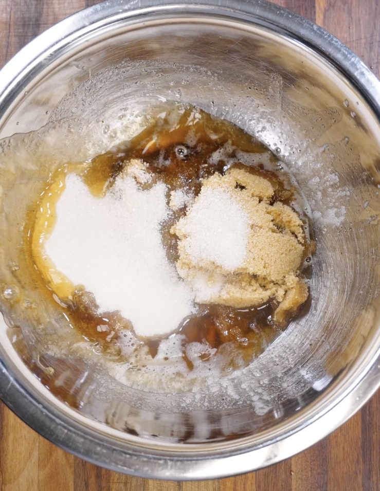 Warm brown butter combined with granulated sugar and dark brown sugar in a stainless steel mixing bowl before whisking to create the cookie dough base.