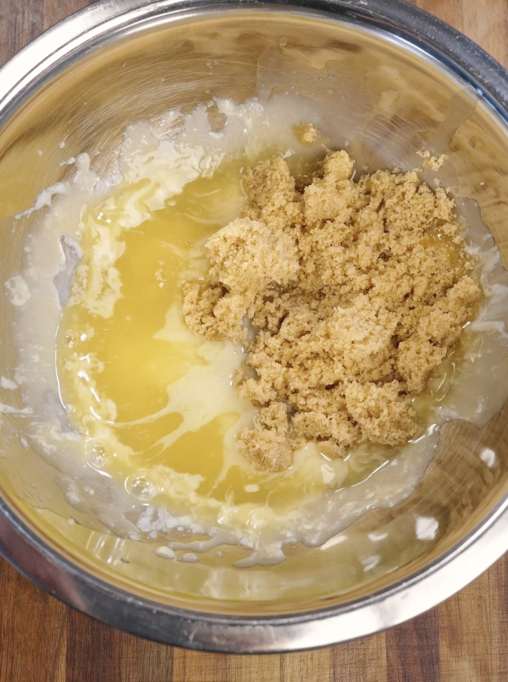 Melted butter combined with granulated sugar and light brown sugar in a mixing bowl before whisking to create a smooth brownie base.