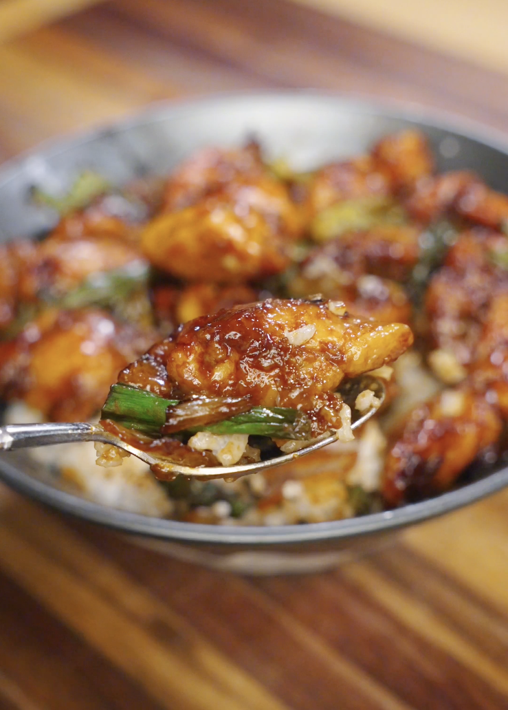 Close up of Mongolian chicken served over white rice, featuring glossy sweet soy glazed chicken thighs, charred onions, and scallions in a shallow bowl.