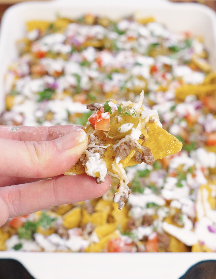A single tortilla chip lifted from sheet pan beef nachos showing layers of seasoned ground beef, melted cheese, pickled jalape&ntilde;os, tomato, onion, and garlic sour cream.