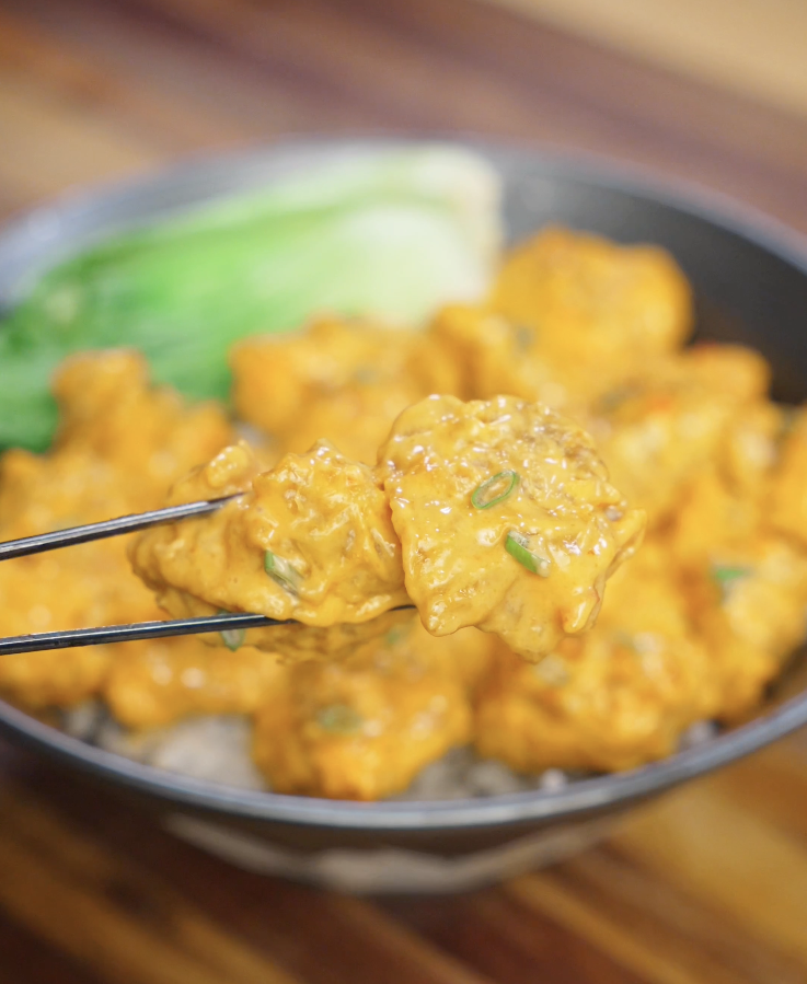 Chopsticks lifting a piece of crispy bang bang chicken coated in creamy sweet chili sauce over a bowl of rice.