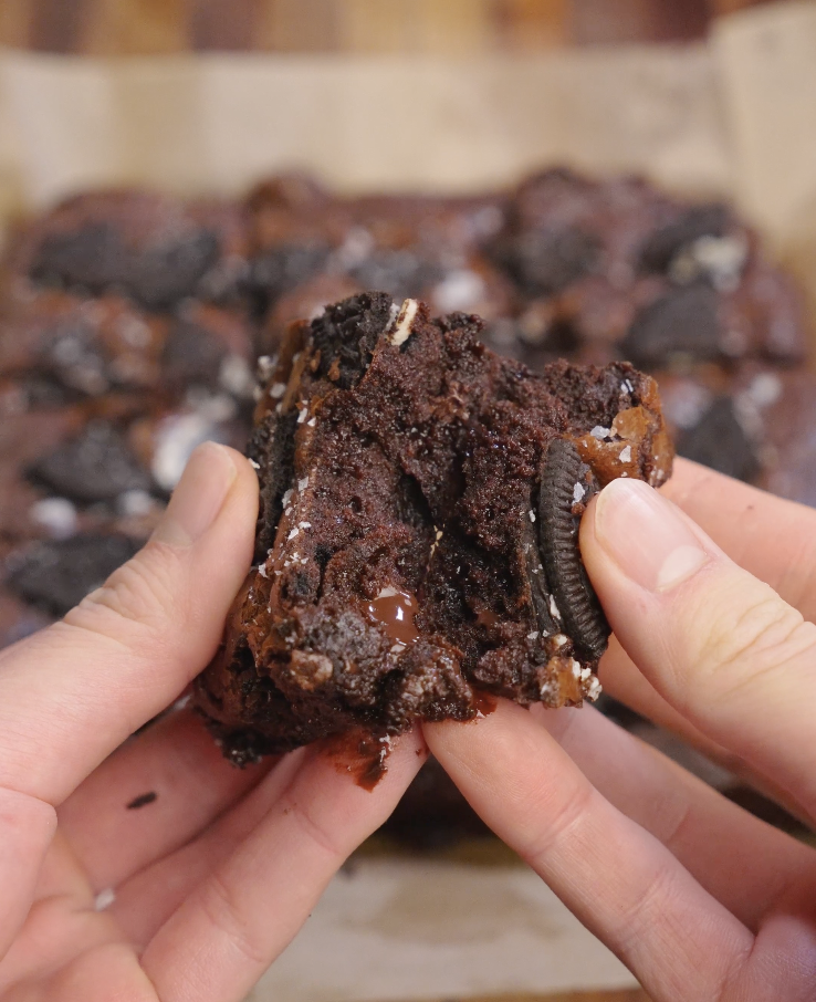 Hands pulling apart a warm fudgy Oreo brownie to reveal a molten center, gooey chocolate pockets, and softened Oreo cookies inside.