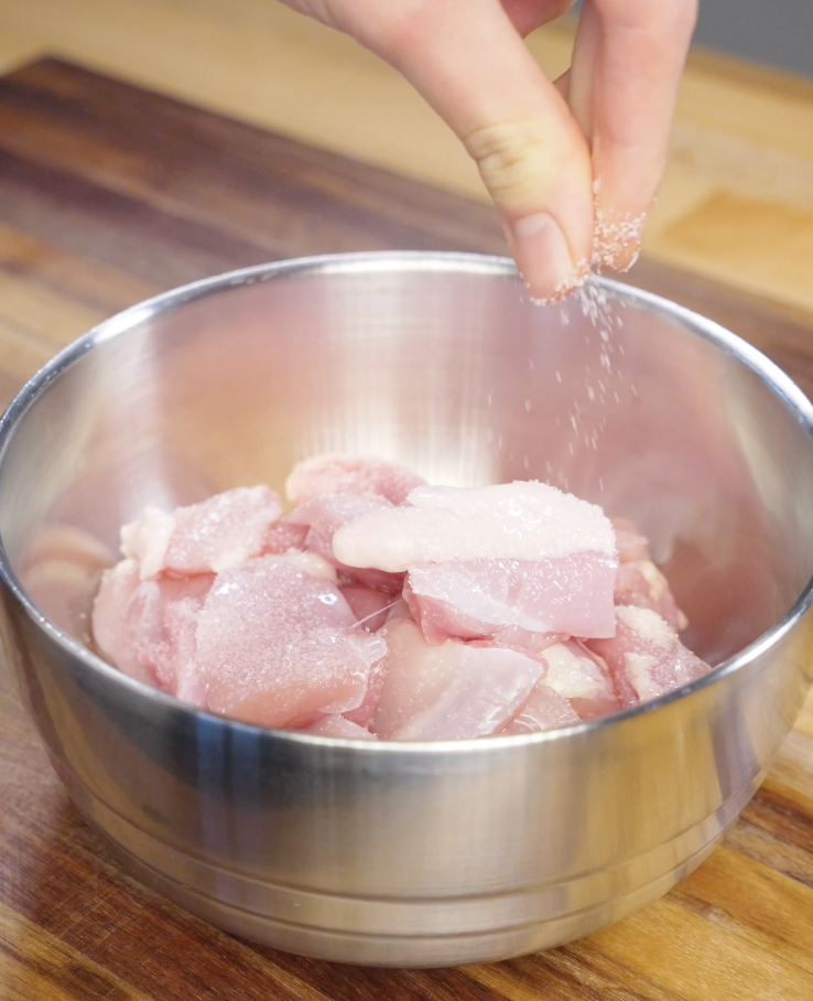 Hand sprinkling salt over bite sized boneless skinless chicken thighs in a metal bowl before marinating for homemade Mongolian chicken.