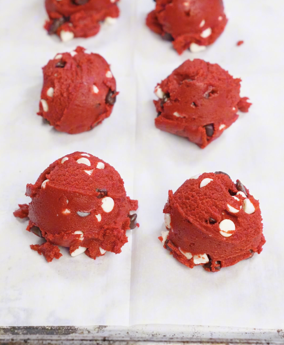 Large scoops of vibrant red velvet cookie dough portioned onto a parchment lined baking sheet for thick, bakery style cookies.