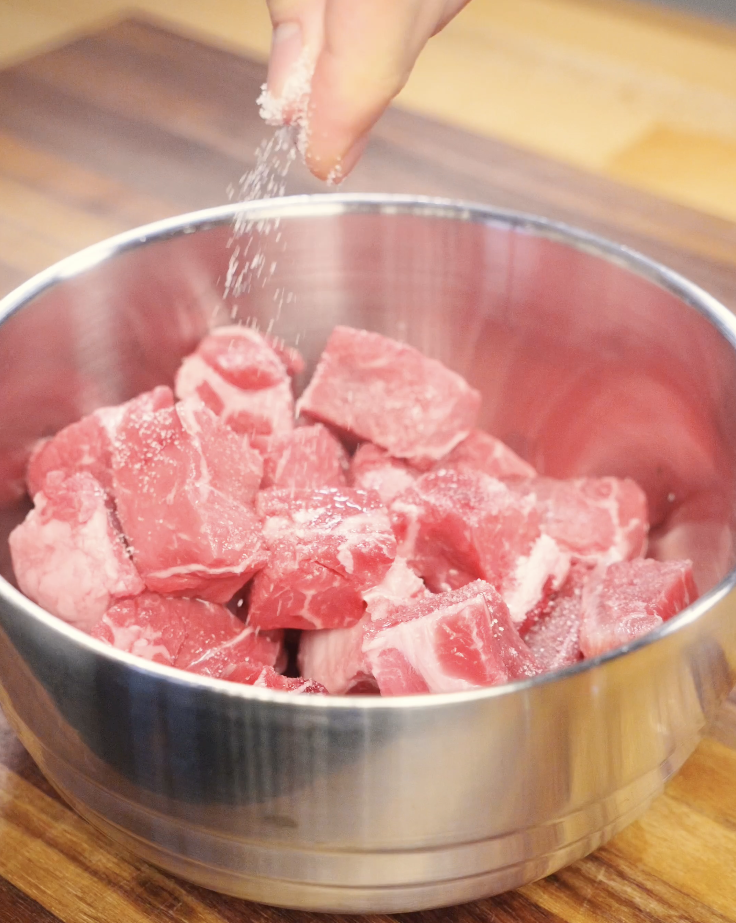 Raw beef cubes in a stainless steel bowl being seasoned with salt before marinating, preparing the beef for a tender and flavorful soy garlic beef recipe.