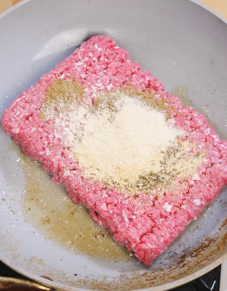 Raw ground beef in a skillet seasoned with salt, onion powder, garlic powder, oregano, and cumin before browning for sheet pan beef nachos.