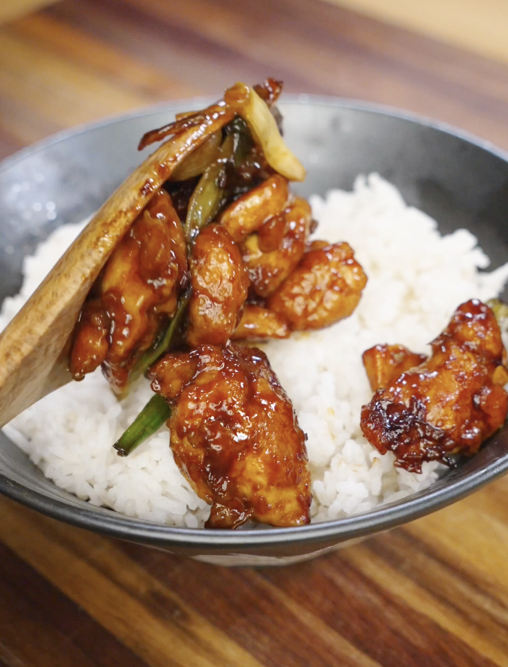 Wooden spoon placing sticky Mongolian chicken with charred onions and scallions over a bowl of steamed white rice for an easy weeknight dinner.